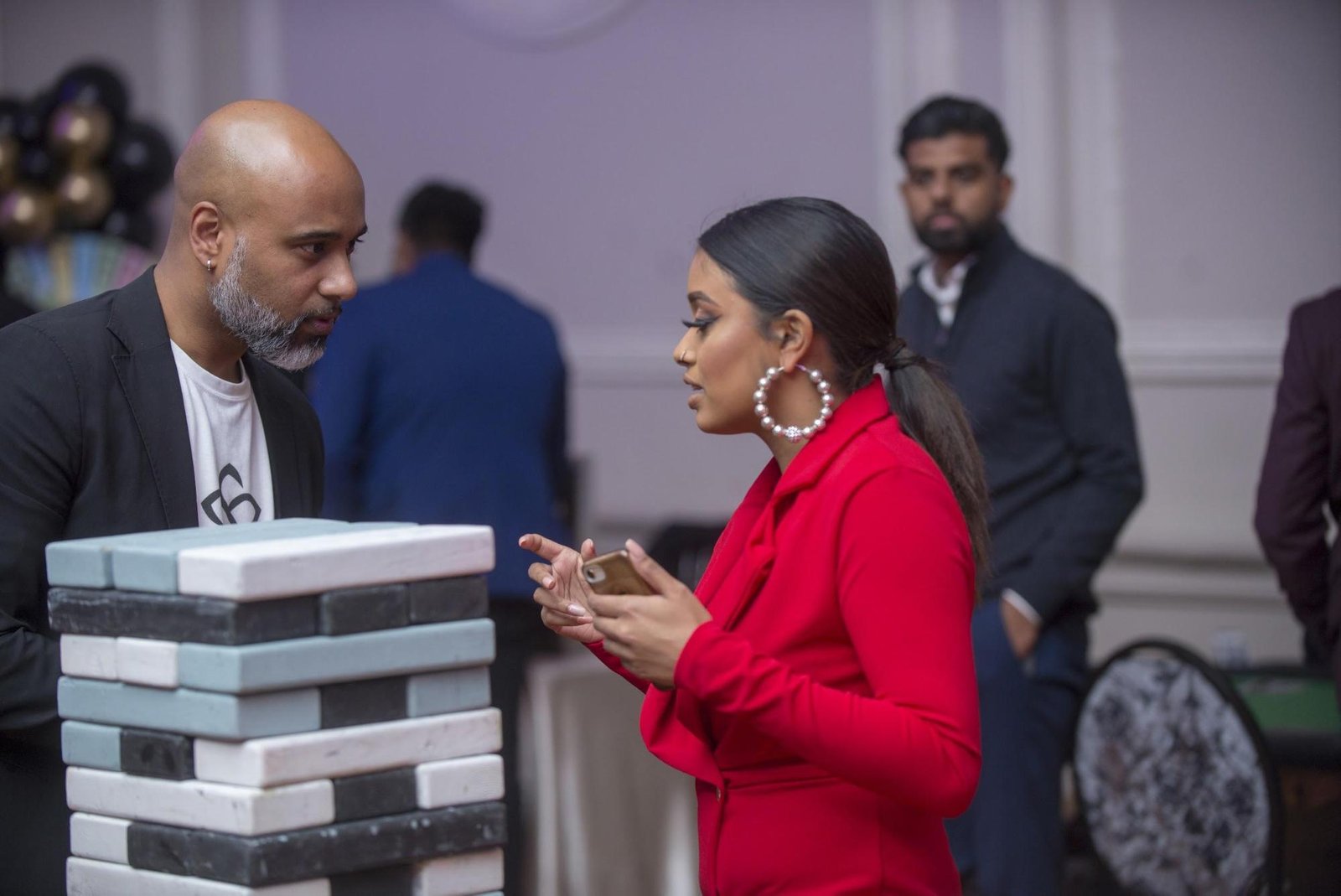 Two individuals engaged in conversation, one in a red blazer and large earrings, discussing while standing near a stack of blocks, with blurred figures in the background, highlighting mentorship and personal development themes.